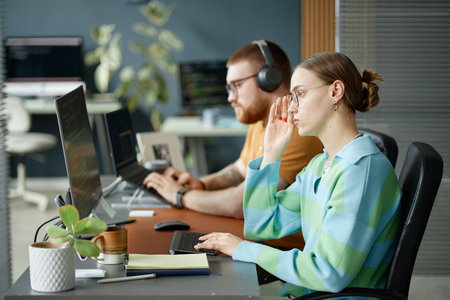 Young woman wearing glasses working at computer in modern office, adjusting eyewear while typing,...