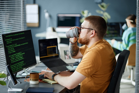 Young adult man with beard and glasses sitting at desk working on computer with code on screen,...