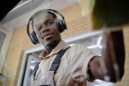 Young adult Black man wearing headphones cleaning glass surface with focused expression, standing...