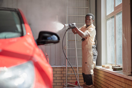 Young adult Black man cleaning red car with high pressure washer indoors, standing near large...