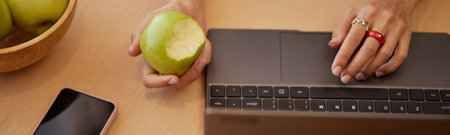 Website header shot of young adult Caucasian woman working on laptop while holding bitten green...