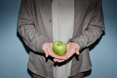 Middle aged man holding green apple in both hands against plain blue background, torso and hands visible, standing upright, face not shown, casual clothingの写真素材