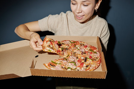 Young adult Caucasian woman smiling while holding open pizza box and lifting slice of pizza, showing excitement and anticipation for eating freshly delivered foodの写真素材