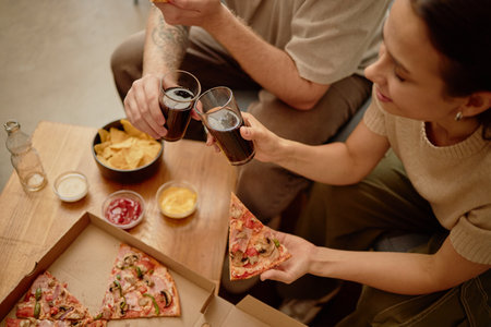 Young adult woman and young adult man sitting together sharing pizza and clinking glasses of soda,...