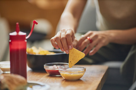 Caucasian young adult woman dipping tortilla chip into cheese sauce while sitting at table with...