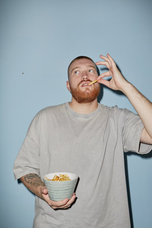 Caucasian young adult man with red beard holding bowl of French fries, eating and looking upward, standing against plain blue background, casual pose with thoughtful expressionの写真素材
