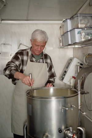 Senior Caucasian man stirring large industrial pot in kitchen, focusing on preparing food, wearing...
