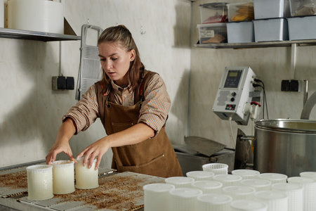 Young adult Caucasian woman working in cheese production facility, arranging cheese molds on...