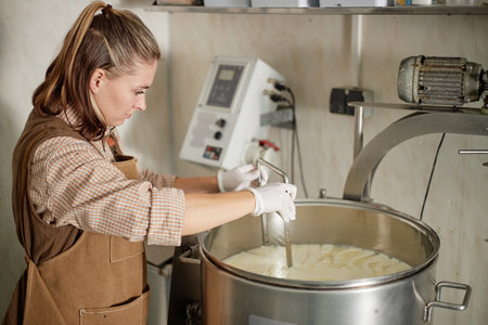 Caucasian young adult woman preparing cheese curds in stainless steel vat using industrial...