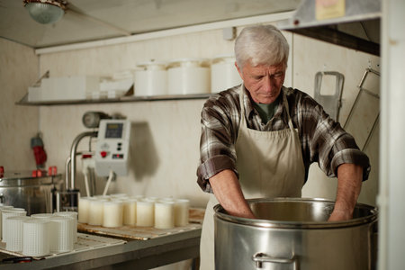 Senior Caucasian man working in cheese production facility, standing at stainless steel vat with...