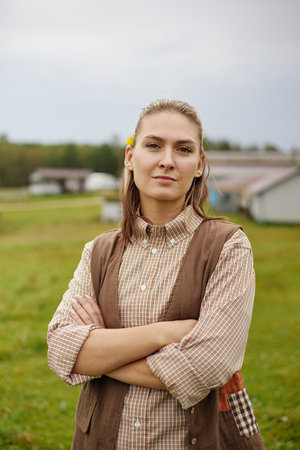Portrait of young adult Caucasian woman standing outdoors with arms crossed, looking confidently at camera, background showing rural landscape with buildings and green fieldの写真素材
