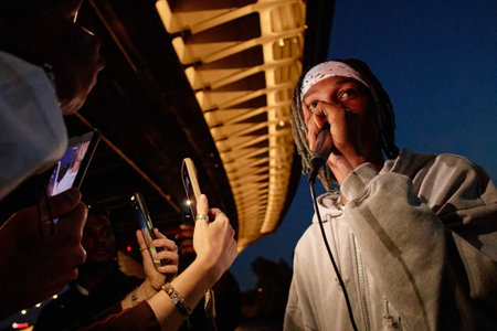 Young Black man performing with microphone outdoors at night, facing crowd holding smartphones,...