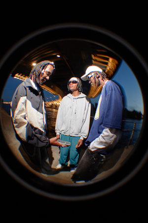 Three Black young adult men standing together under bridge at night, one holding smartphone while others watching, all wearing casual clothing and sunglasses, urban settingの写真素材