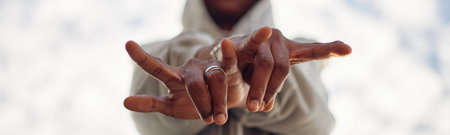 Header of Black young adult man extending hands toward camera making expressive gesture, focus on fingers and ring, blurred face in background, outdoor setting with sky visibleの写真素材