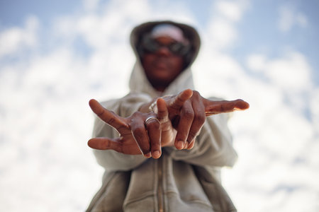Black young adult man wearing hoodie standing outdoors extending both hands toward camera making...