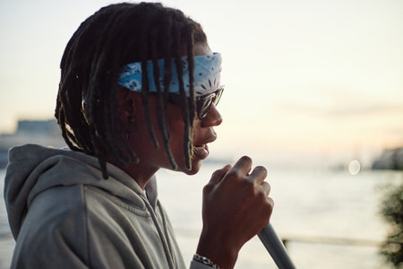 Black man wearing sunglasses and bandana holding microphone, performing outdoors near waterfront...