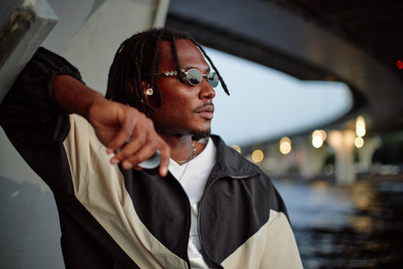 Black young adult man wearing sunglasses leaning against urban structure under bridge looking away from camera with thoughtful expression outdoorsの写真素材