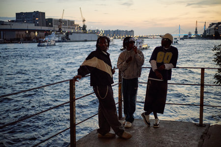 Three Black men standing by waterfront railing during sunset, two engaging in conversation while third looking away, cityscape and industrial cranes visible in backgroundの写真素材