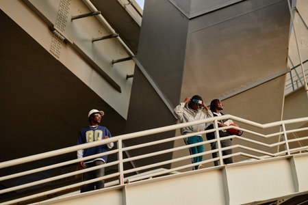 Three Black men standing on outdoor bridge, two holding basketball and adjusting hats while looking away, urban setting with metal structure in background, casual postureの写真素材