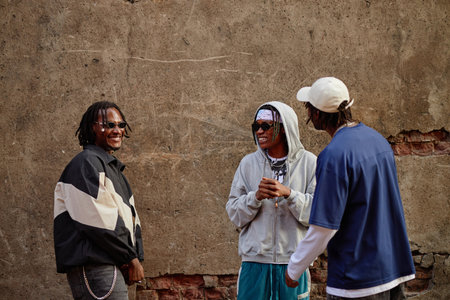 Three Black young adult men standing outdoors laughing and talking together near textured concrete...