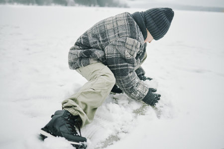 Caucasian boy crouching on snowy ground shaping snow with hands outdoors during winter, wearing warm clothing and knit hat, focusing on activityの写真素材