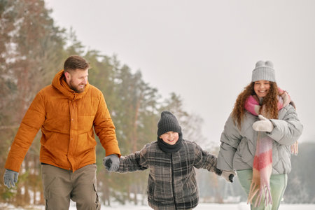 Caucasian man, woman and child walking together outdoors in winter, holding hands and smiling, snow covered trees in backgroundの写真素材