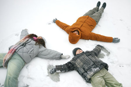 Three people lying on snow making snow angels, including man, woman, and child, all dressed in winter clothing, arms and legs spread out, enjoying winter activity outdoorsの写真素材