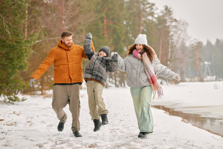 Caucasian man, woman, and boy walking together in snowy forest, parents holding hands of smiling...