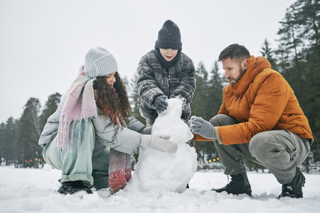 Man, woman, and child building snowman together outdoors in winter forest, all wearing warm clothing, collaborating and shaping snow, trees visible in backgroundの写真素材