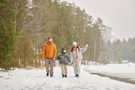 Caucasian man, woman, and boy walking together on snowy path near forest and lake, holding hands and talking, winter landscapeの写真素材