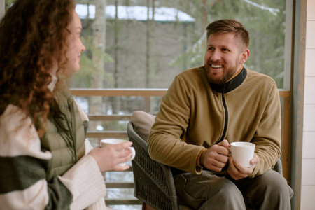 Caucasian young adult man smiling and talking while holding coffee cup, sitting outdoors with Caucasian young adult woman, both engaging in relaxed conversation on porchの写真素材