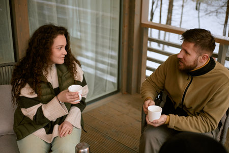 Caucasian young woman and man sitting on balcony holding mugs, engaging in conversation, winter...