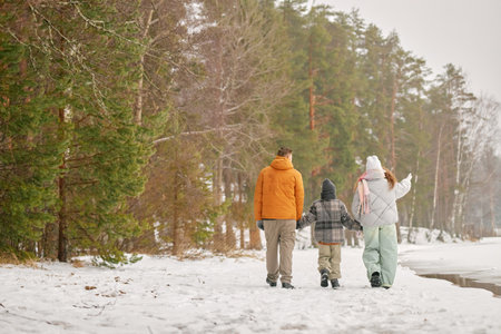 Man, woman, and child walking together holding hands along snowy forest path near frozen lake, backs facing camera, winter trees surrounding groupの写真素材