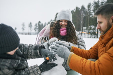 Caucasian man, woman and child building snowman together outdoors in winter landscape, all wearing winter clothing and smiling while shaping snow figureの写真素材