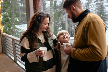 Caucasian woman, man, and child standing together on outdoor wooden terrace holding mugs, child...