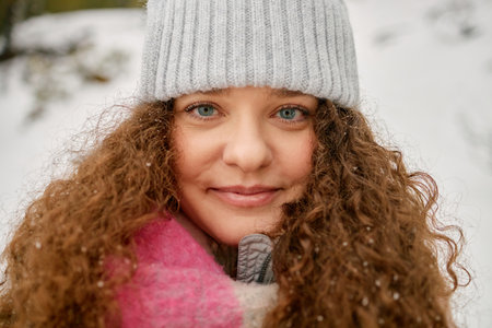 Portrait of young adult Caucasian woman smiling and looking into camera outdoors in snowy winter setting, long curly hair visible under knit hat, snowflakes resting on hairの写真素材