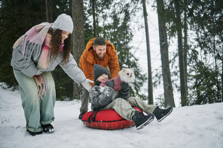 Man, woman, and child playing together outdoors in snowy forest, man and woman pushing smiling child sitting on snow tube holding small fluffy dogの写真素材