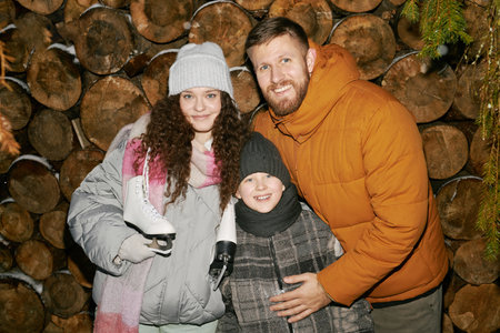 Portrait of Caucasian man, woman holding ice skates, and child boy smiling together in winter clothing in front of stacked firewoodの写真素材