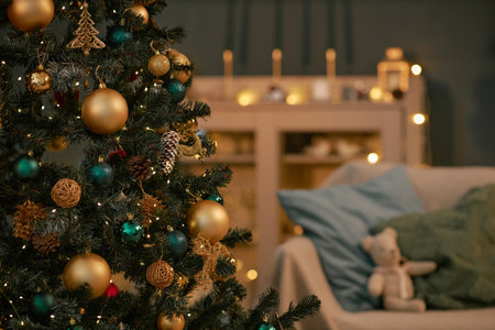 Christmas tree decorated with gold and teal ornaments standing in living room with blurred background showing sofa, festive lights illuminating branches and decorationsの写真素材