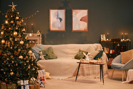 Christmas tree decorated with ornaments and lights standing beside sofa in cozy living room, wrapped gifts arranged under tree, candles and framed art on wall creating festive settingの写真素材