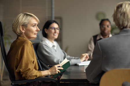 Caucasian middle aged woman holding notebook participating in business meeting with multiethnic group of women sitting around table discussing project in office settingの写真素材