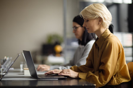 Caucasian middle aged woman working on laptop at desk in modern office, sitting beside young adult woman with dark hair, both focused on computer tasks in professional environmentの写真素材