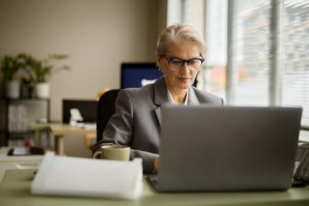 Senior Caucasian woman working on laptop in modern office, wearing eyeglasses and formal suit, concentrating on screen, sitting at desk with coffee mug and documents, daylight through windowの写真素材