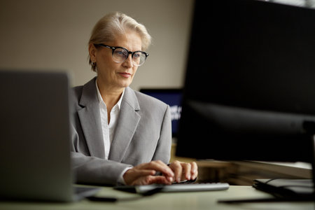 Senior Caucasian woman wearing eyeglasses working at computer in modern office, typing on keyboard and focusing on monitor, demonstrating professional business environmentの写真素材