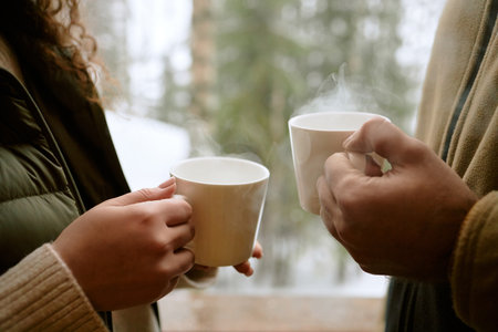 Woman and man holding steaming mugs outdoors, standing close together with hands visible, sharing...