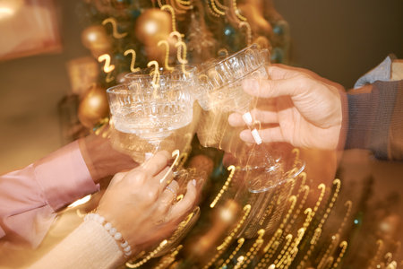 Men and women clinking crystal champagne glasses during festive celebration, hands visible in foreground with blurred golden holiday decorations in backgroundの写真素材
