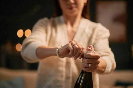 Caucasian young adult woman opening champagne bottle with both hands, standing indoors with blurred background, focus on hands and bottle, face partially visibleの写真素材