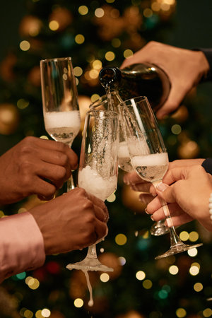 Diverse group of young adult hands holding champagne glasses clinking together while sparkling wine being poured, celebrating festive occasion in front of decorated treeの写真素材