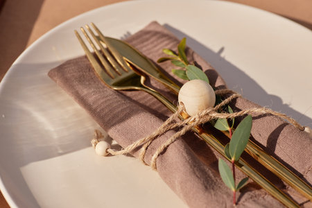 Closeup showing gold fork and knife placed on folded napkin tied with twine and wooden bead,...