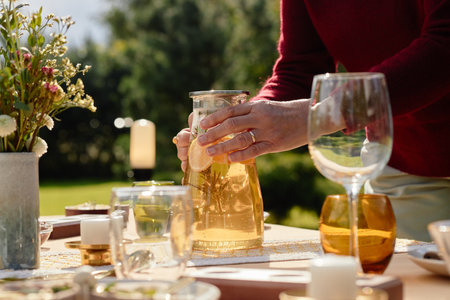 Middle aged Caucasian woman arranging outdoor table, pouring beverage from glass pitcher among...